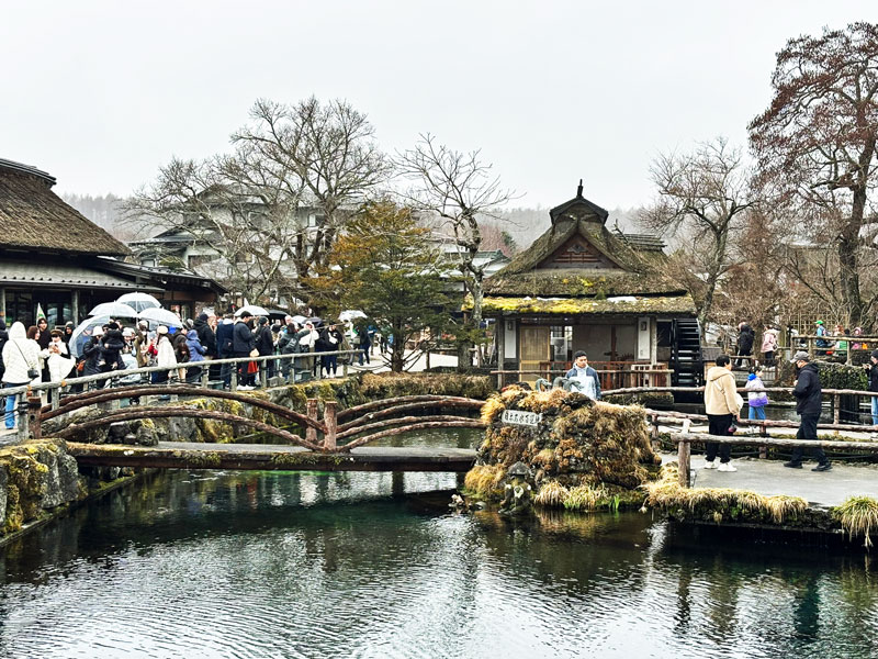 A scenic view of a traditional Japanese village with a wooden bridge crossing a small pond. People with umbrellas are seen walking along the bridge and gathering around the area, surrounded by trees and traditional wooden buildings.