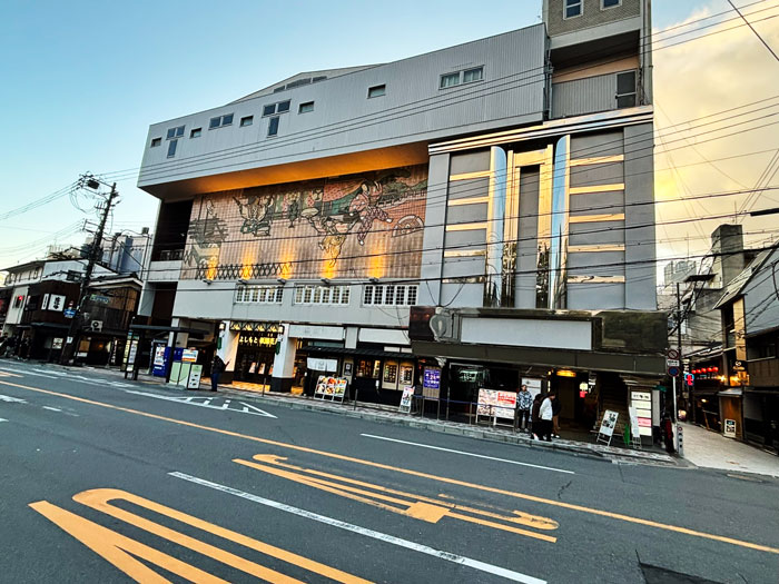 Building with a modern facade featuring a large mural, located on a street in Kyoto, Japan, with light reflections and power lines in the foreground.