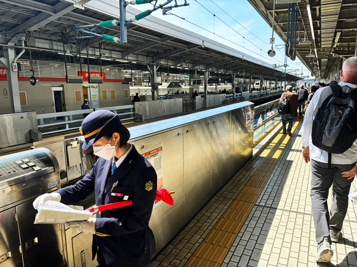 A train station scene in Osaka, featuring a station attendant in uniform checking a schedule while passengers walk by on the platform.