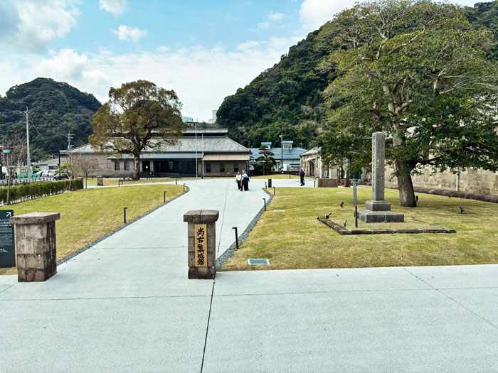 A view of Sengan-en, featuring grassy pathways, historical buildings, trees, and stone monuments, with mountains in the background.