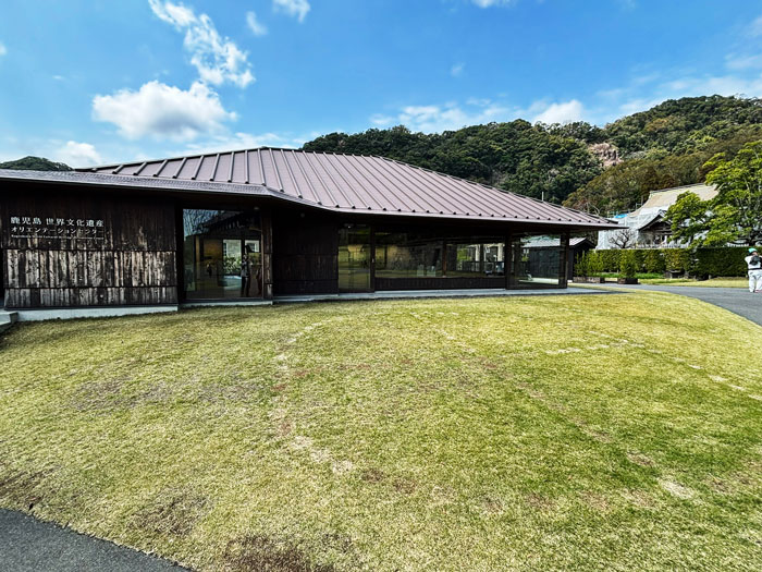 A modern wooden building with a sloped roof, surrounded by a grassy area and hills in the background.