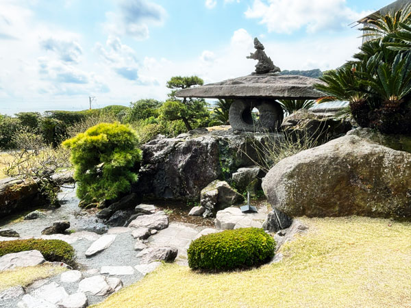A traditional Japanese garden featuring a stone lantern, lush greenery, and a small stream flowing over rocks, under a partly cloudy sky.