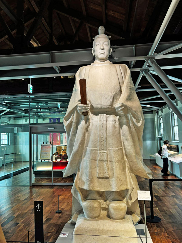A large statue of a historical figure dressed in traditional attire, holding a wooden tablet, displayed in a museum setting.