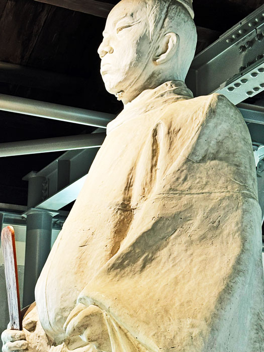 A close-up of a statue depicting a historical figure, dressed in traditional clothing, holding a sword, set against a modern architectural background.