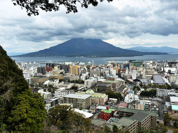 A panoramic view of a city with buildings and a waterfront, featuring a prominent mountain in the background under a cloudy sky.